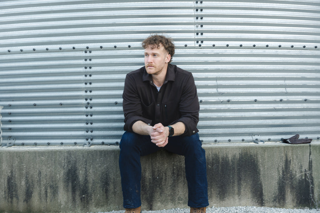 Man sitting on a concrete ledge against a metal shutter wearing Barbell Jeans and Flannel.