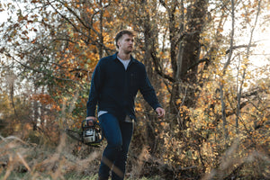 Man in barbell flannel and jeans walking in woods with chainsaw