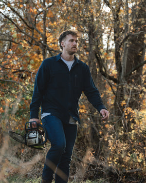 Man in barbell flannel and jeans walking in woods with chainsaw