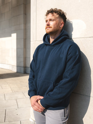 Man wearing a Cadet Foundation Barbell hoodie standing against a light-colored wall.