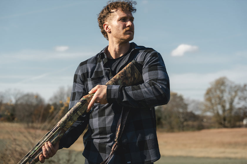 Man holding a shotgun in an outdoor setting with trees and sky in the background in Barbell Frontier Flannel